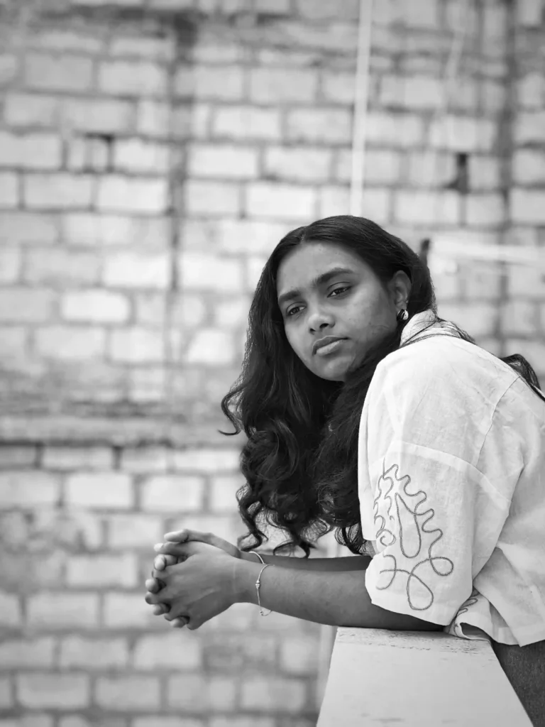 This black-and-white photo shows a freelance digital marketer in Bangalore wearing jeans and a shirt, standing and leaning slightly against a wall. She is a little hunched, with her hands locked together. Her hair is untied, and she looks like she is thinking about something, creating a calm and thoughtful mood.