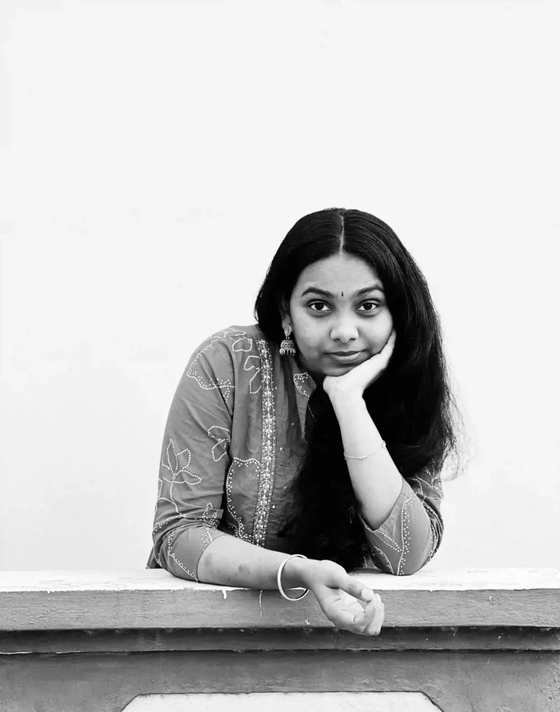 This black-and-white photo shows a freelance digital marketer in Bangalore wearing a churidar, leaning against a wall with her hair untied, gently touching her chin with her left hand, while wearing a bangle on her right hand and jhumka earrings